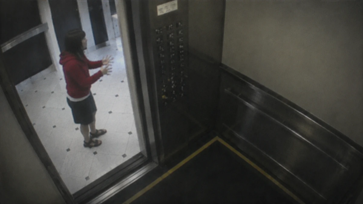 Dramatised recreation of the elevator surveillance footage: a young woman in a red hoodie stands in the tiled hallway outside the open elevator doors, both hands raised in front of her as if gesturing to someone unseen