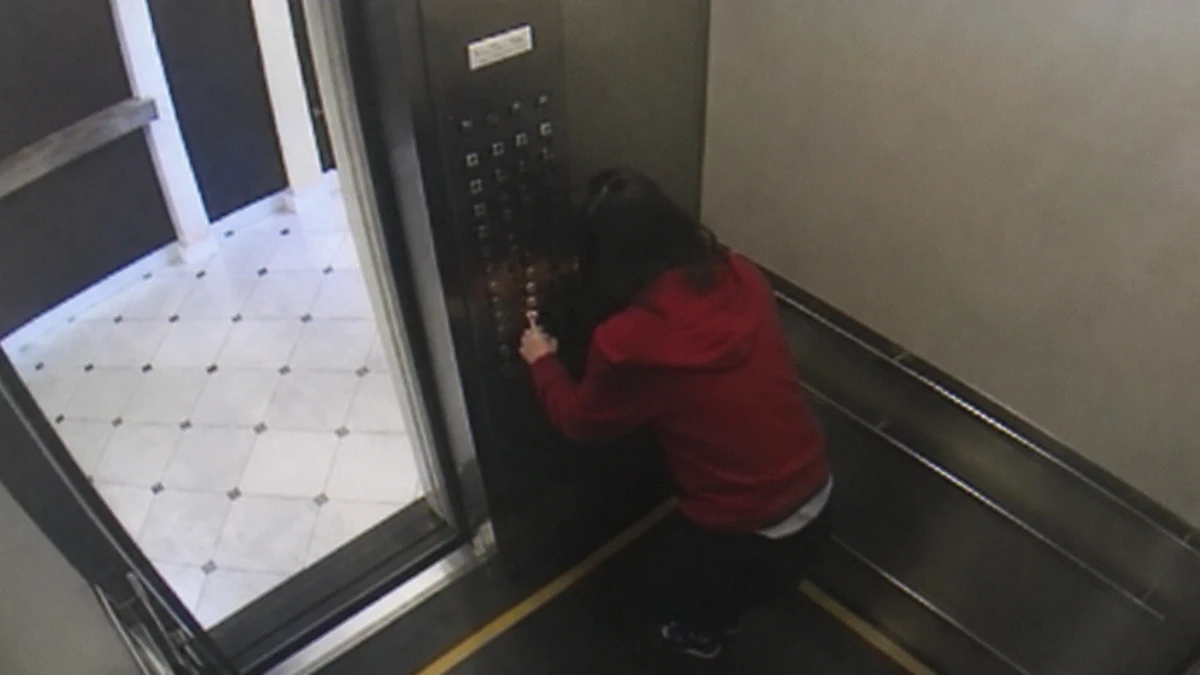 Dramatised recreation of the Cecil Hotel elevator surveillance footage: seen from the ceiling camera, a young woman in a red hoodie bends close to the elevator button panel and presses the buttons while the doors stand open onto a tiled corridor